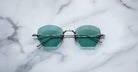 A pair of Jacques Marie Mage Oatman hexagonal rimless sunglasses in teal with thin metallic arms, displayed on a white marble surface.
