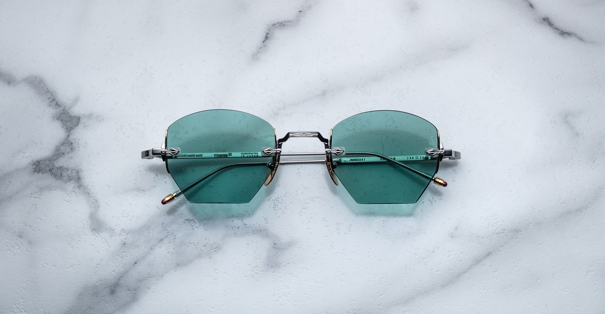 A pair of Jacques Marie Mage Oatman hexagonal rimless sunglasses in teal with thin metallic arms, displayed on a white marble surface.
