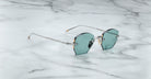A pair of Jacques Marie Mage Oatman round rimless sunglasses with blue-green tinted lenses and thin metal arms rests on a white marble surface.