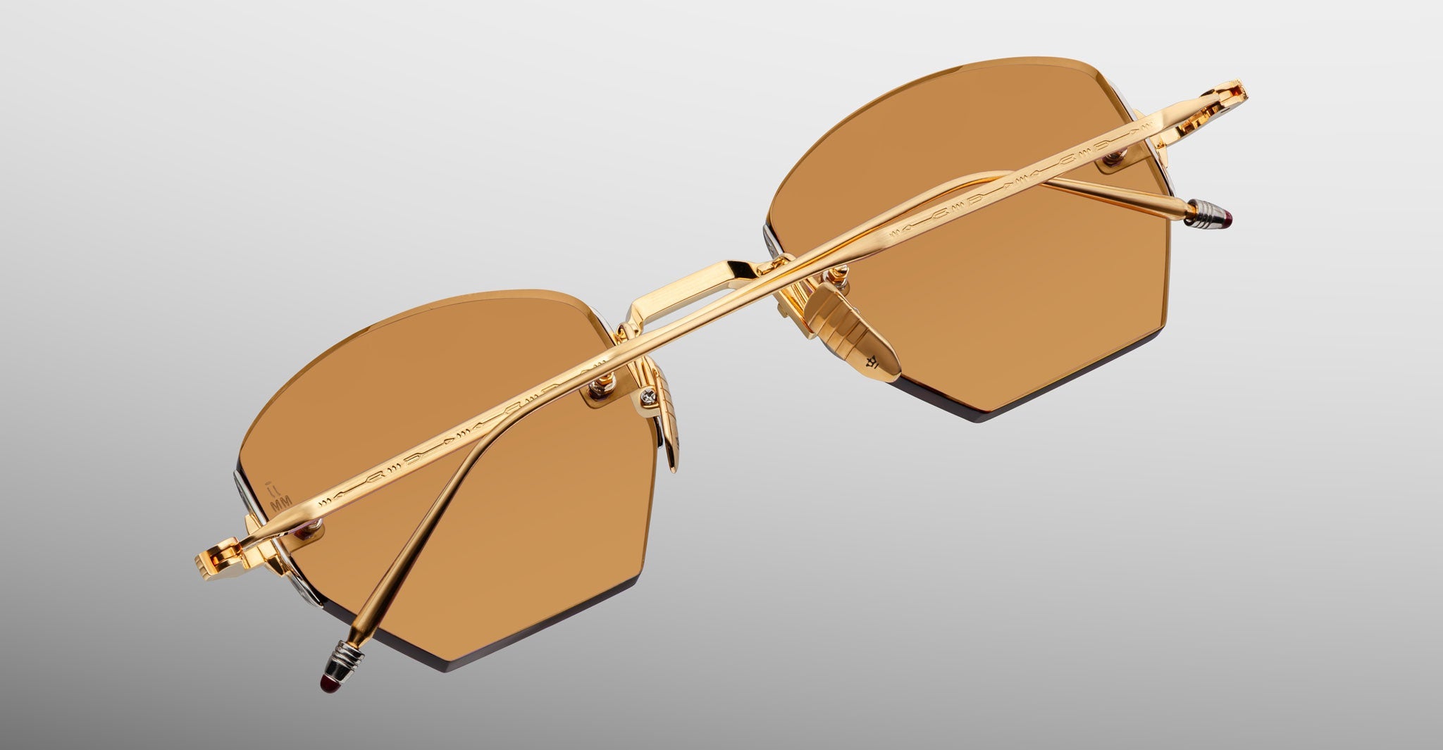 Jacques Marie Mage Oatman sunglasses by Jacques Marie Mage, featuring gold frames and rimless hexagonal amber-tinted lenses with a geometric design, are showcased on a light gray background, seen from the back.