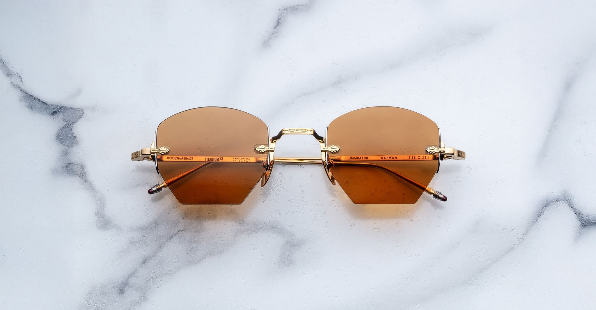 A pair of Jacques Marie Mage Oatman sunglasses with brown-tinted rimless hexagonal lenses and gold frames rests elegantly on a white marble surface.