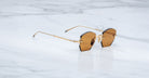 The Jacques Marie Mage Oatman sunglasses feature gold hexagonal rimless frames and brown-tinted geometric lenses, inspired by handmade Japanese eyewear, displayed on a white marble surface.