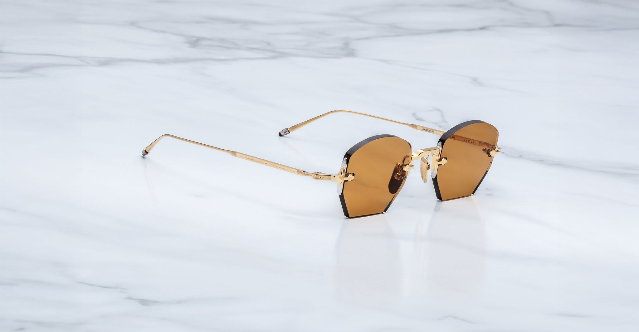 The Jacques Marie Mage Oatman sunglasses feature gold hexagonal rimless frames and brown-tinted geometric lenses, inspired by handmade Japanese eyewear, displayed on a white marble surface.