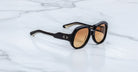 The Jacques Marie Mage Ocean Child sunglasses, featuring black thick frames, brown gradient lenses, and silver dot accents near the hinges, rest on a white marble surface.