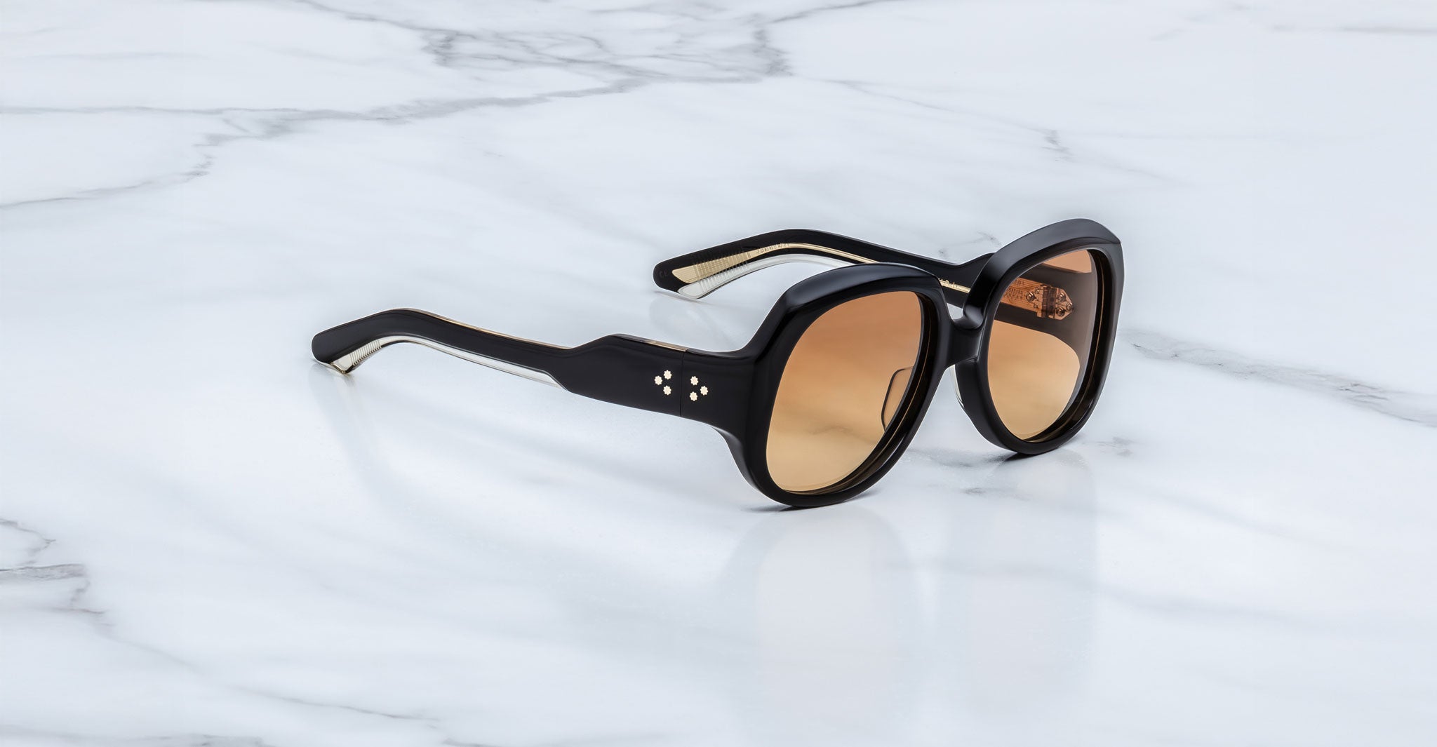 The Jacques Marie Mage Ocean Child sunglasses, featuring black thick frames, brown gradient lenses, and silver dot accents near the hinges, rest on a white marble surface.