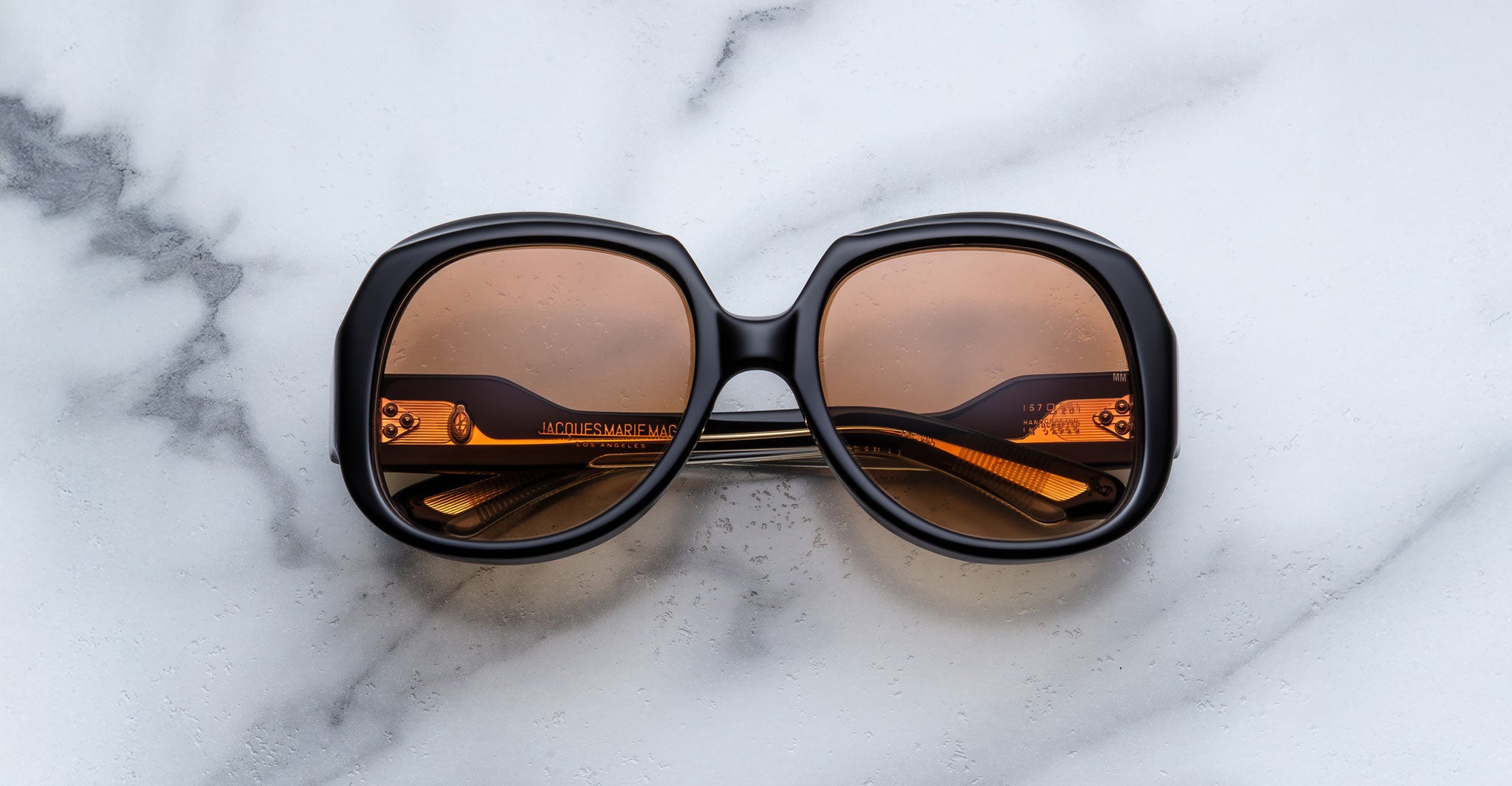 The Jacques Marie Mage Ocean Child, large round sunglasses with dark frames and amber-tinted lenses, are folded on a white marble surface with gray veining.