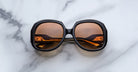 The Jacques Marie Mage Ocean Child, large round sunglasses with dark frames and amber-tinted lenses, are folded on a white marble surface with gray veining.