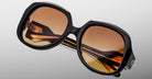 Close-up of Jacques Marie Mage Ocean Child oversized sunglasses, featuring thick black frames and brown gradient lenses, folded on a light gray background.
