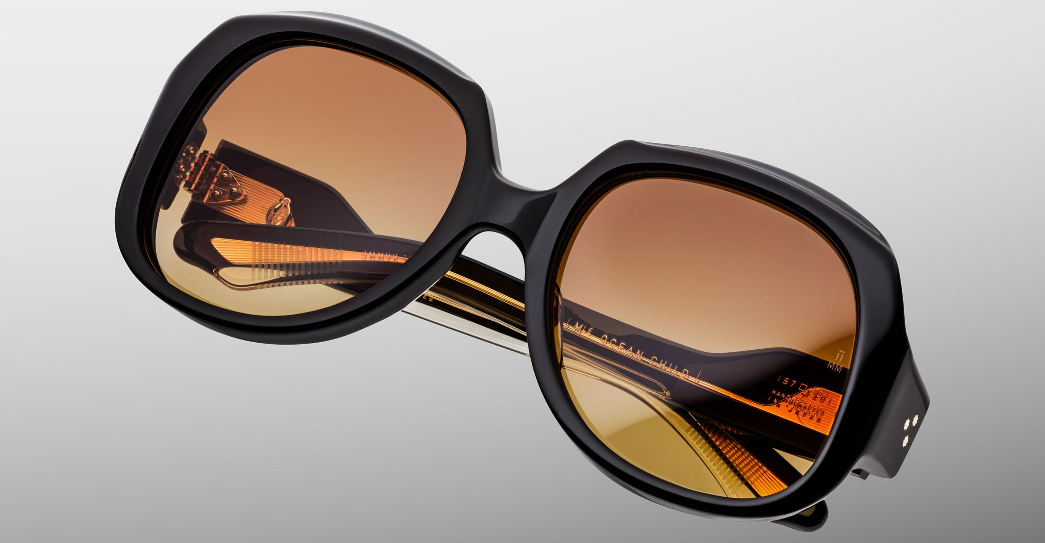 Close-up of Jacques Marie Mage Ocean Child oversized sunglasses, featuring thick black frames and brown gradient lenses, folded on a light gray background.