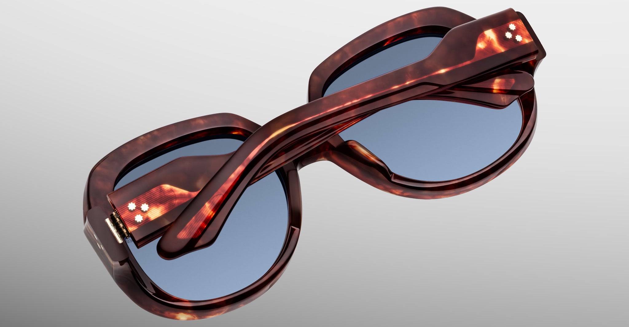 The Jacques Marie Mage Ocean Child sunglasses, in tortoiseshell with blue-tinted lenses, are folded neatly with crossed arms and displayed on a light gray background.
