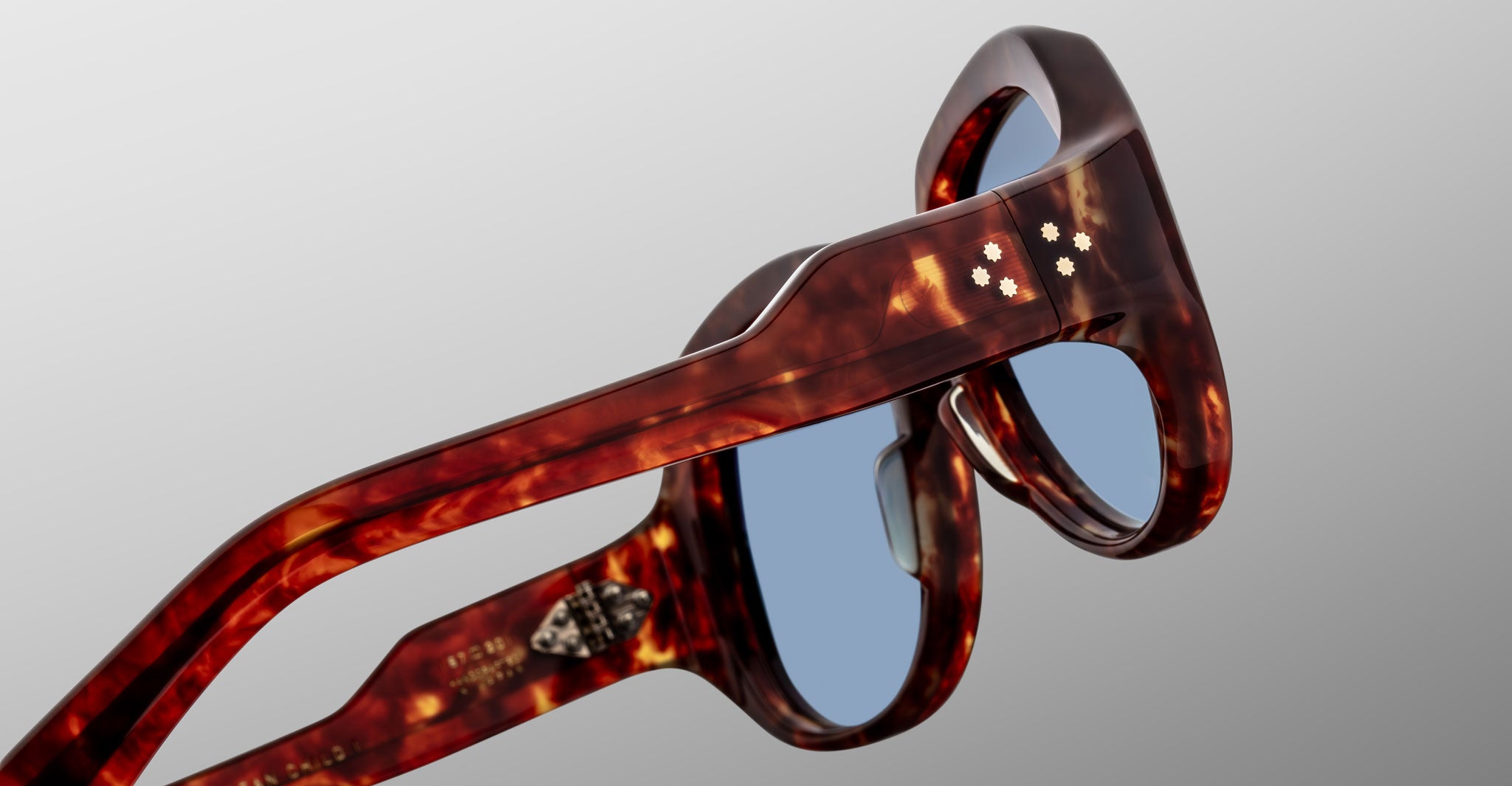 Close-up of Jacques Marie Mage Ocean Child sunglasses in brown tortoiseshell with blue lenses, highlighting the temples and decorative hinges against a plain grey background.