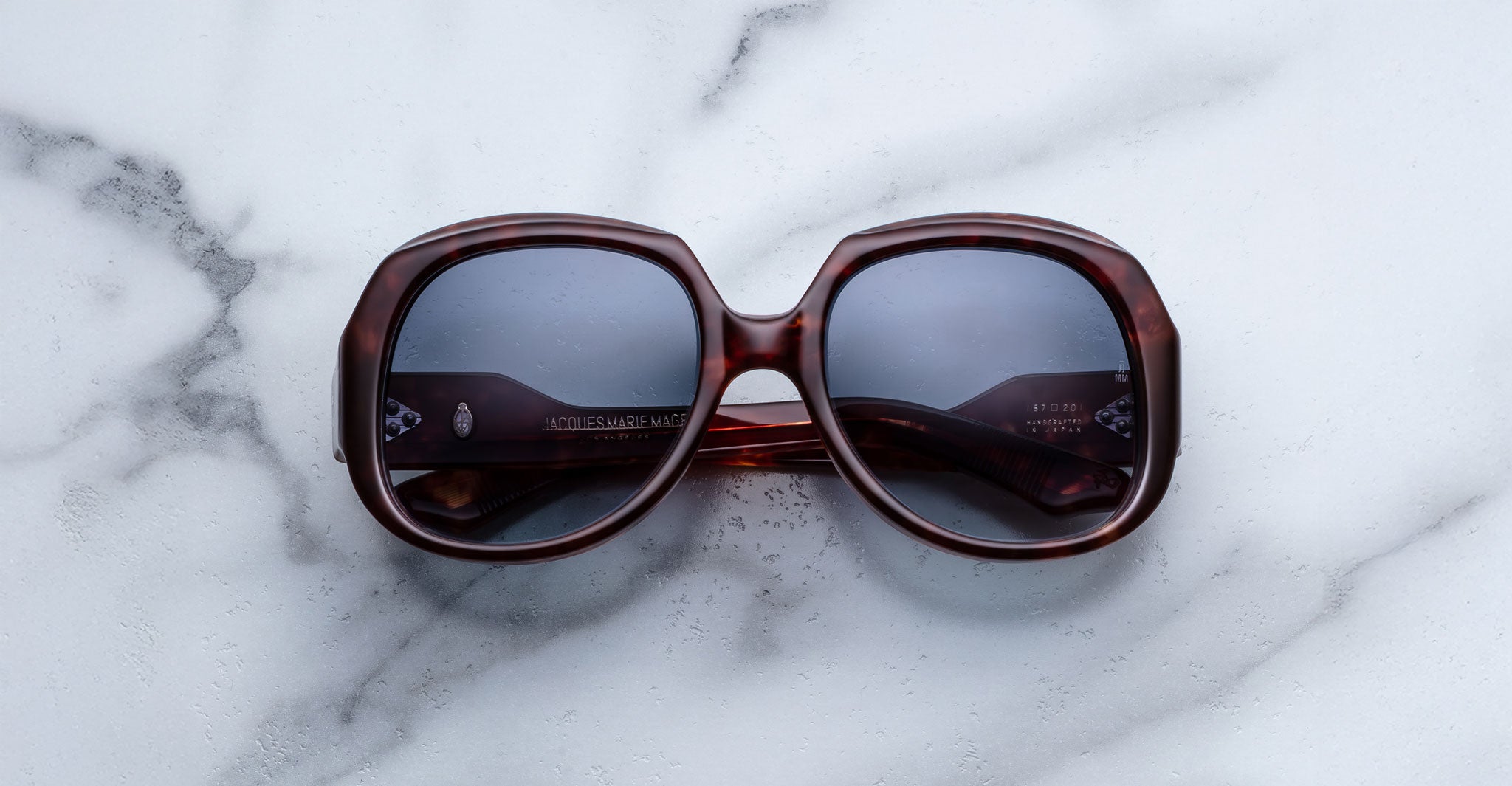 The Jacques Marie Mage Ocean Child sunglasses, featuring an oversized square design in dark brown with tinted lenses, rest on a white marble surface.