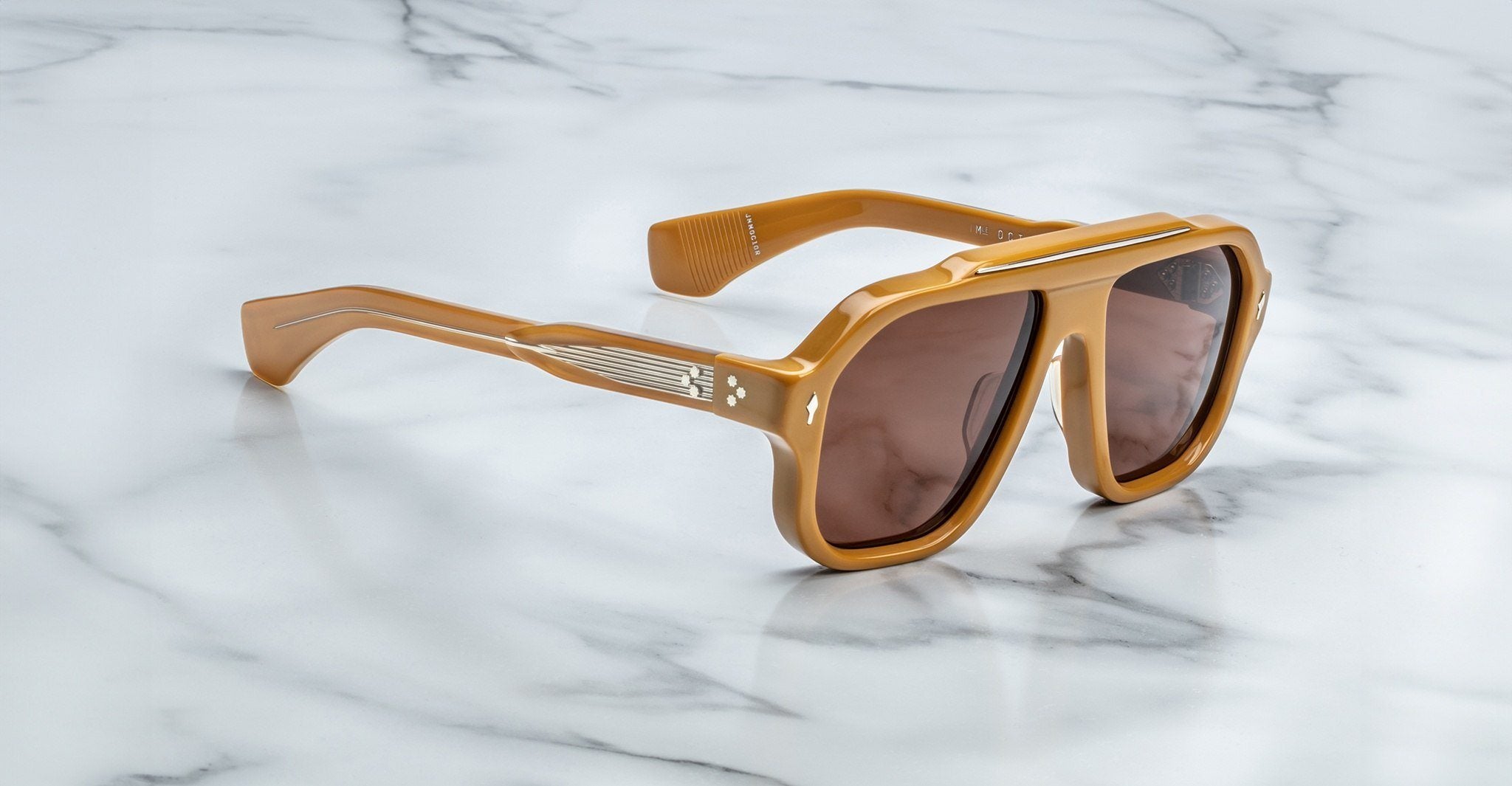 The Jacques Marie Mage Octavian sunglasses, featuring oversized brown frames with tinted lenses, thick angular design, and metallic accents on the arms, rest on a polished white marble surface.