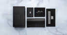 A flat lay of five black Jacques Marie Mage Cash items on white marble: a textured box, a small booklet, a card with white signature, a large case or wallet, and a black card with white text.