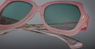 Close-up of Jacques Marie Mage Perreti sunglasses with pink frames and green-tinted lenses, highlighting detailed design on transparent, curved arms against a neutral gray background.
