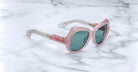 The Jacques Marie Mage Perreti sunglasses, featuring a pink cat-eye frame, teal lenses, and gold arms, rest on a white marble surface.