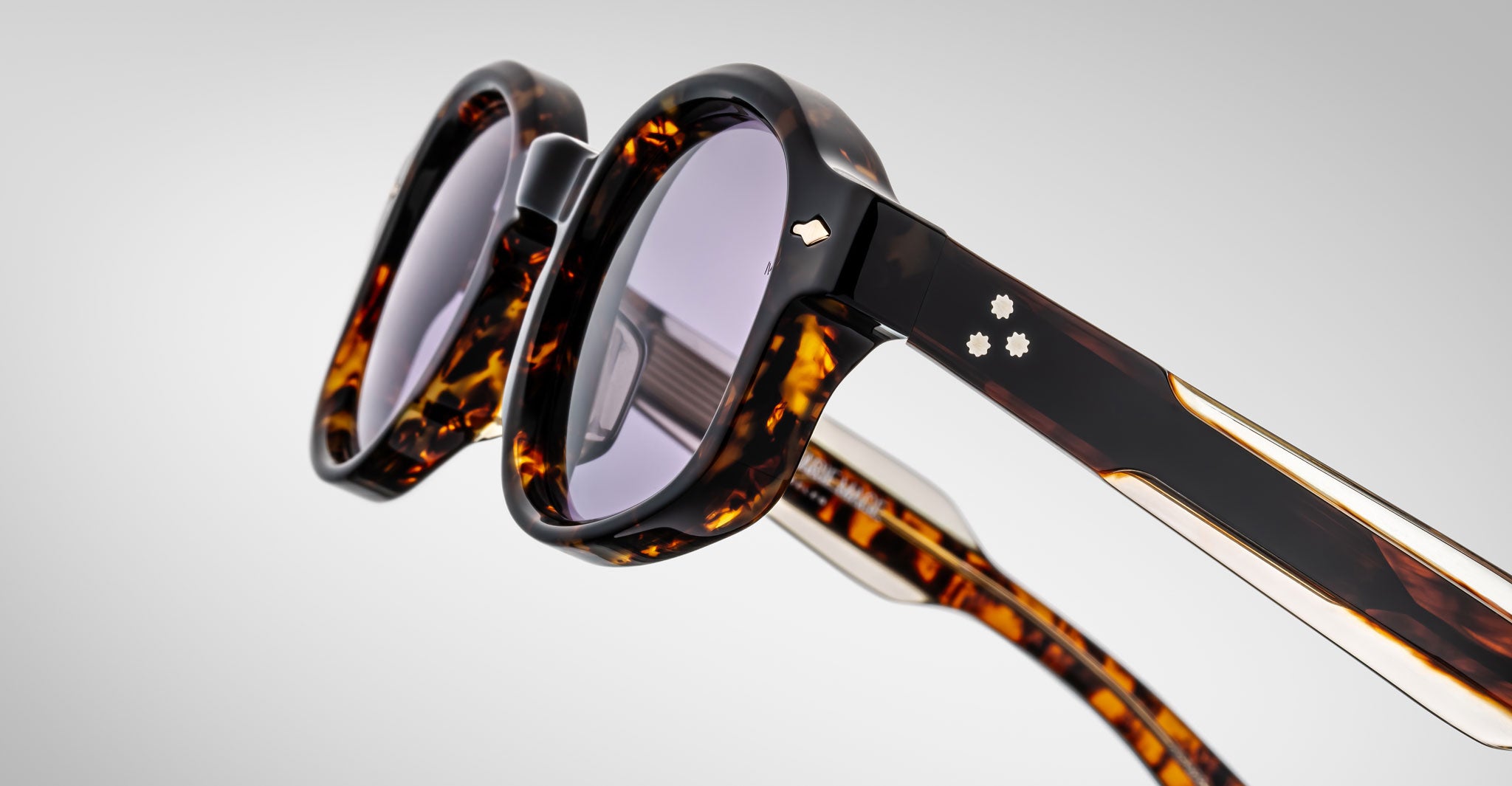 Close-up of Jacques Marie Mage Petrus tortoiseshell sunglasses with thick frames and purple-tinted lenses. Temples feature two small decorative metal flowers and a gold accent. Set against a plain light gray background.