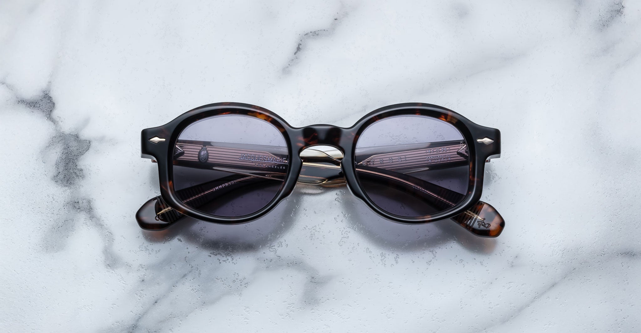 Jacques Marie Mage Petrus sunglasses, featuring round dark lenses and a tortoiseshell frame, are folded and set on a white marble surface.