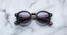Jacques Marie Mage Petrus sunglasses, featuring round dark lenses and a tortoiseshell frame, are folded and set on a white marble surface.