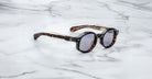 The Jacques Marie Mage Petrus round sunglasses with a tortoiseshell frame and dark lenses rest on a white marble surface, photographed from the side.