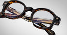 Close-up of Jacques Marie Mage Petrus tortoiseshell round eyeglasses with clear lenses, highlighting the detailed texture and branding on the temples, set against a plain light background.