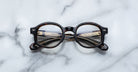 The Jacques Marie Mage Petrus eyeglasses, with round dark tortoise-shell frames and patterned arms, rest folded on a white marble surface, their details visible through the lenses.