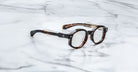 Jacques Marie Mage Petrus round eyeglasses with thick tortoiseshell acetate frames are displayed open on a white marble surface with grey veining.