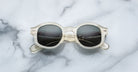 The Jacques Marie Mage Petrus sunglasses, featuring round clear frames and dark lenses, are displayed on a white marble surface with gray veining.