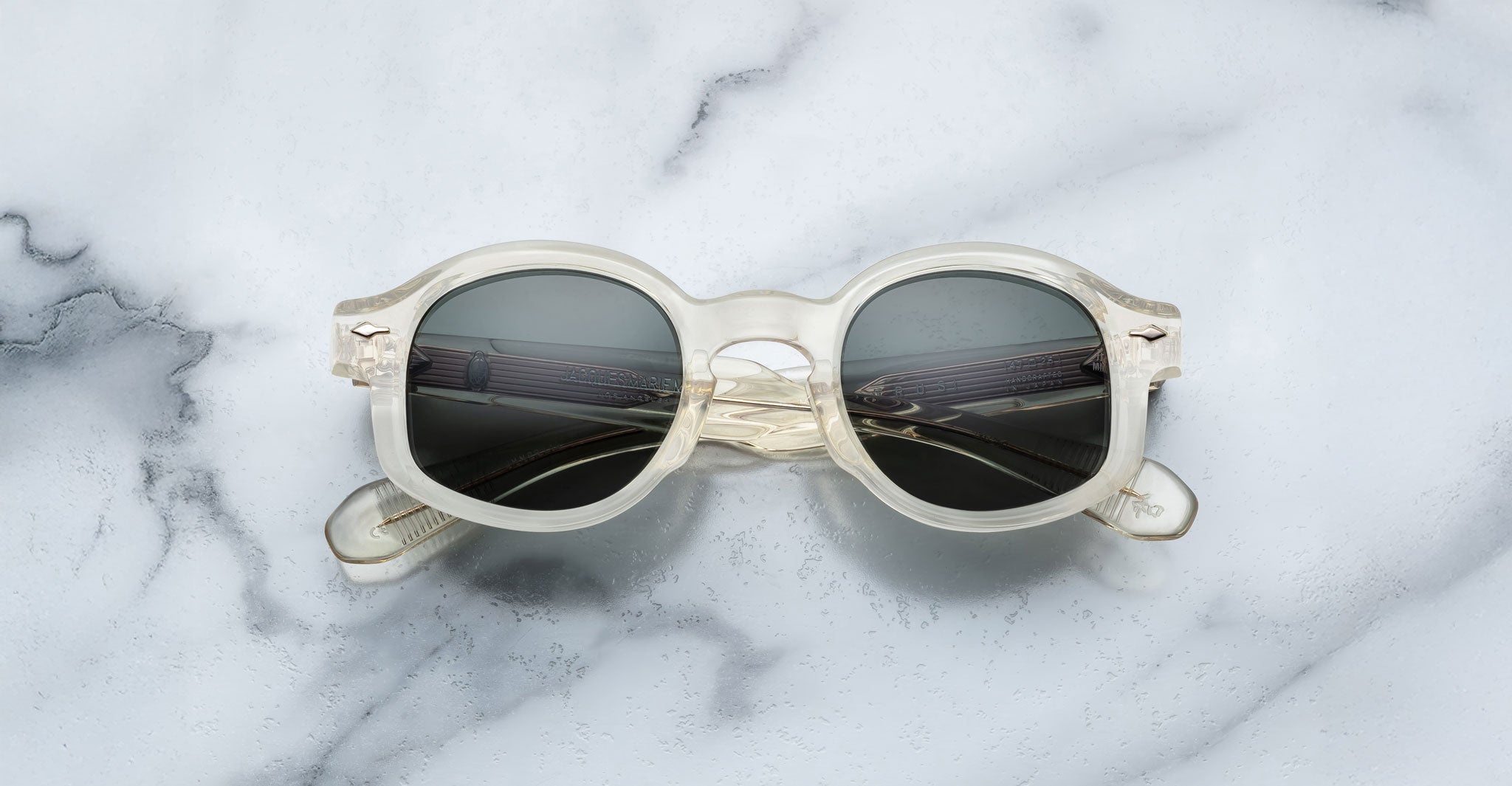 The Jacques Marie Mage Petrus sunglasses, featuring round clear frames and dark lenses, are displayed on a white marble surface with gray veining.