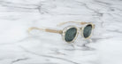 Jacques Marie Mage Petrus sunglasses with clear, round frames and dark lenses are displayed on a white marble surface. The transparent design features subtle beige tones and visible hinges on the temples.