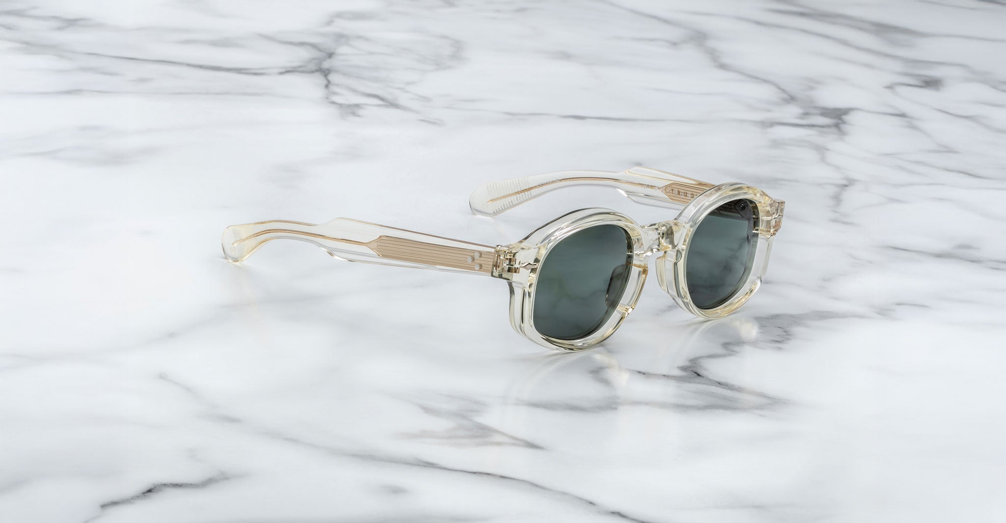 Jacques Marie Mage Petrus sunglasses with clear, round frames and dark lenses are displayed on a white marble surface. The transparent design features subtle beige tones and visible hinges on the temples.