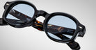 A close-up of Jacques Marie Mage Petrus eyeglasses with black frames and blue-tinted lenses, highlighting detailed engravings and text inside the tortoiseshell-patterned arms.