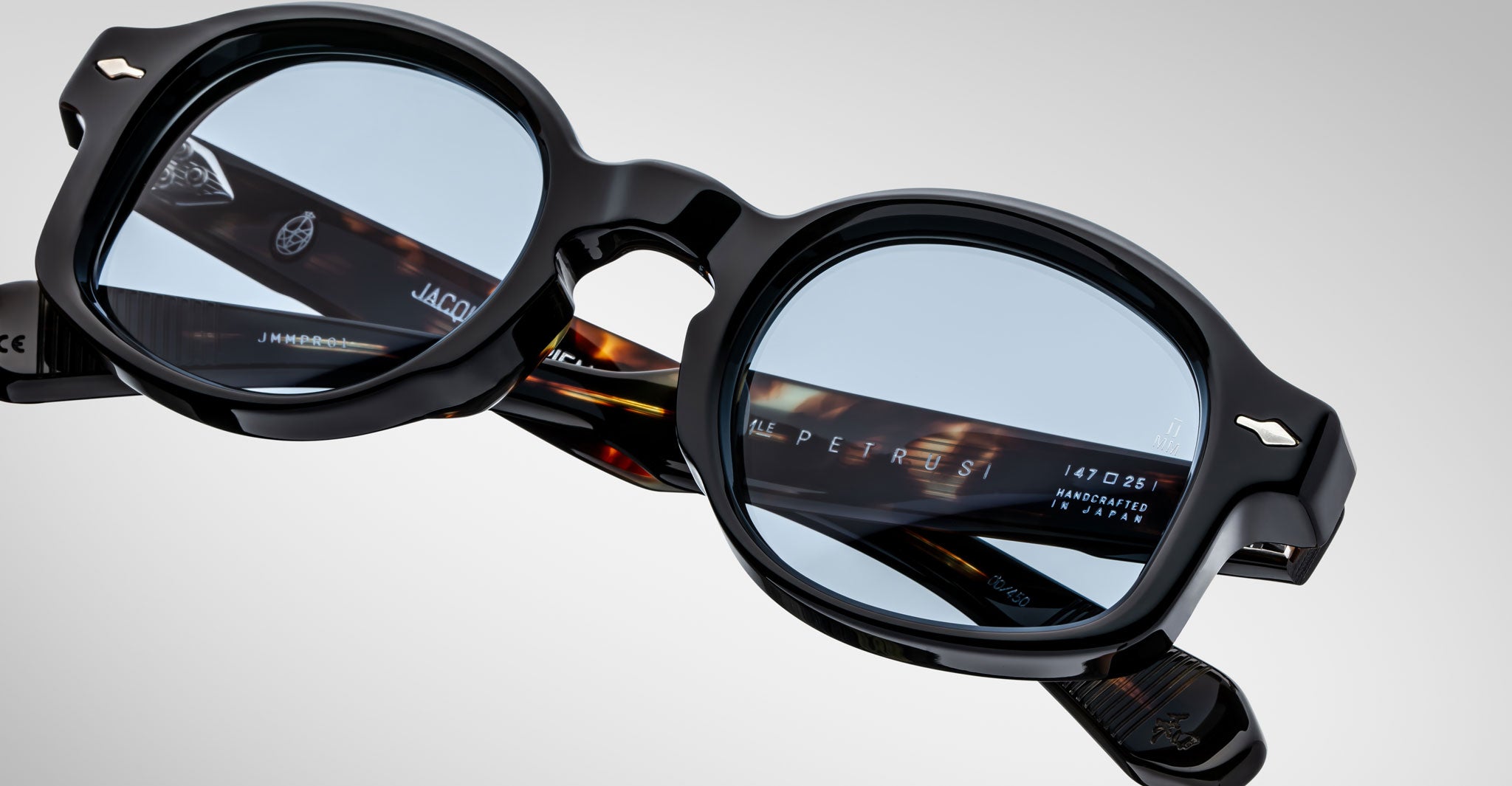 A close-up of Jacques Marie Mage Petrus eyeglasses with black frames and blue-tinted lenses, highlighting detailed engravings and text inside the tortoiseshell-patterned arms.