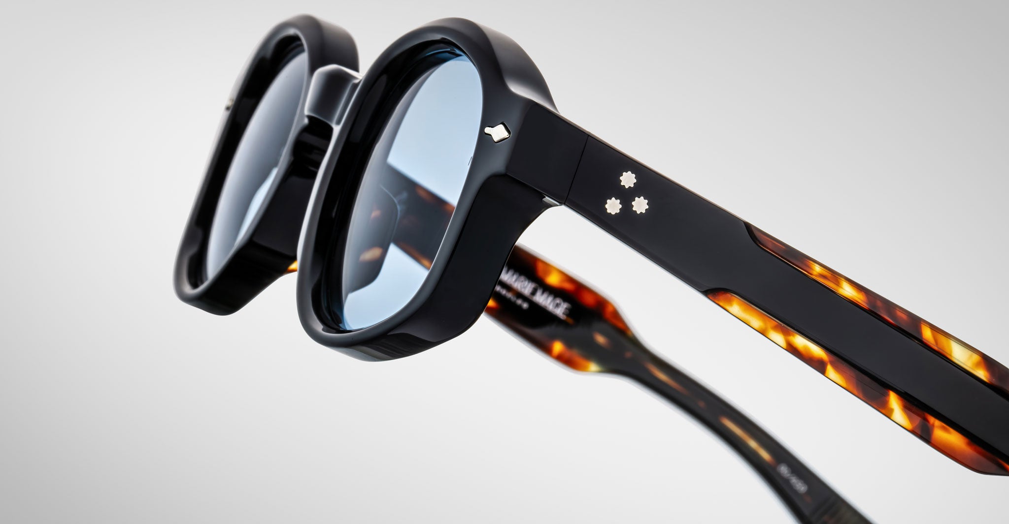 Close-up of Jacques Marie Mage Petrus sunglasses featuring blue lenses, black frames, and tortoiseshell-patterned arms on a light gray background. Small silver decorative accents appear on both the frames and arms.