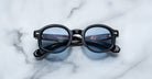 The Jacques Marie Mage Petrus round black eyeglasses with blue-tinted lenses rest on a white marble surface, showing a reflection of the inside of the folded arms and some small text in the lenses.