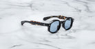 Jacques Marie Mage Petrus round sunglasses with black frames and blue-tinted lenses rest on a white marble surface. The arms feature a subtle tortoiseshell pattern.
