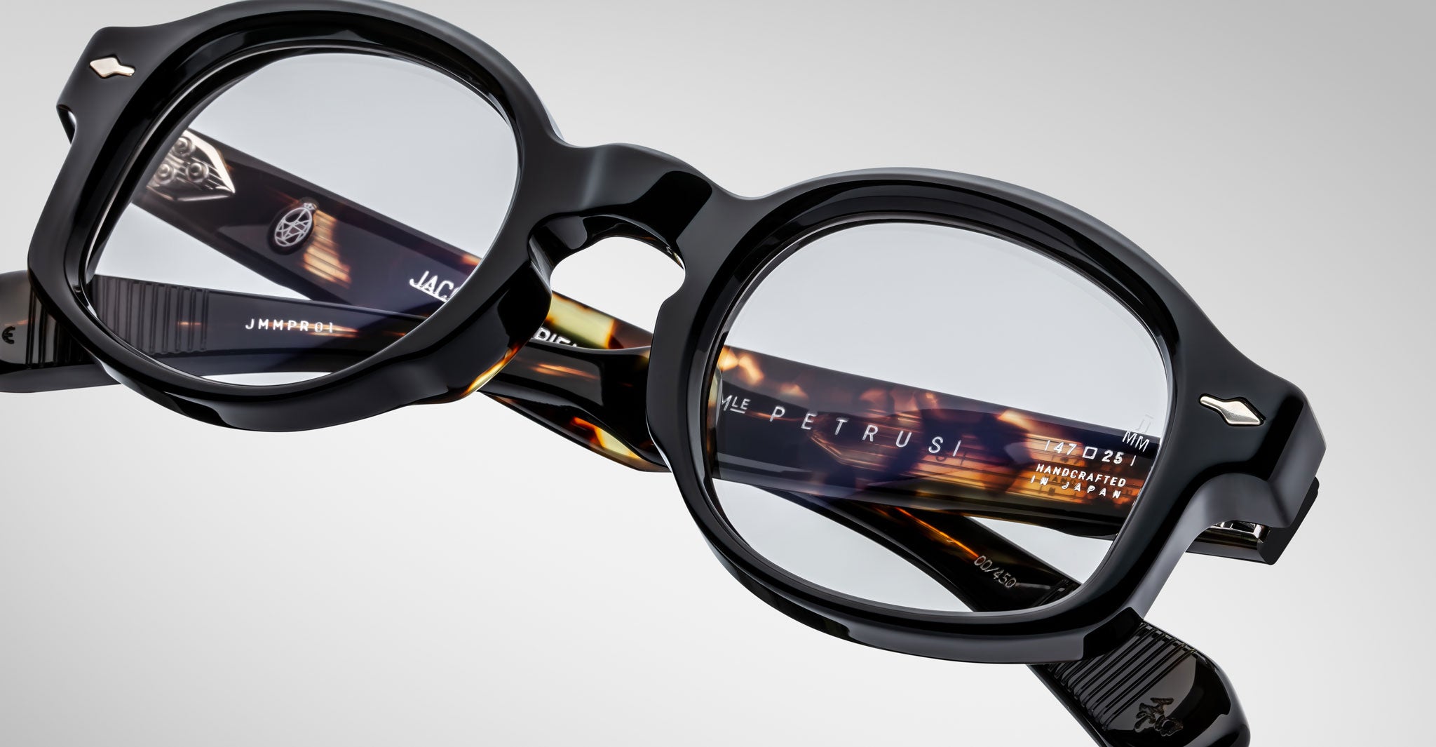 Close-up of Jacques Marie Mage Petrus black round eyeglasses with thick frames. The inside arms feature a tortoiseshell pattern and branded text, shown against a plain, light background.