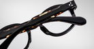 Close-up of two Jacques Marie Mage Petrus eyeglasses with thick, glossy frames—one in black, one in tortoiseshell—stacked atop each other against a plain light background.