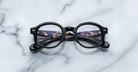 A pair of Jacques Marie Mage Petrus round black eyeglasses lies folded on a white marble surface. The inner arms, partially visible, reveal text and catch a faint reflection of lights.