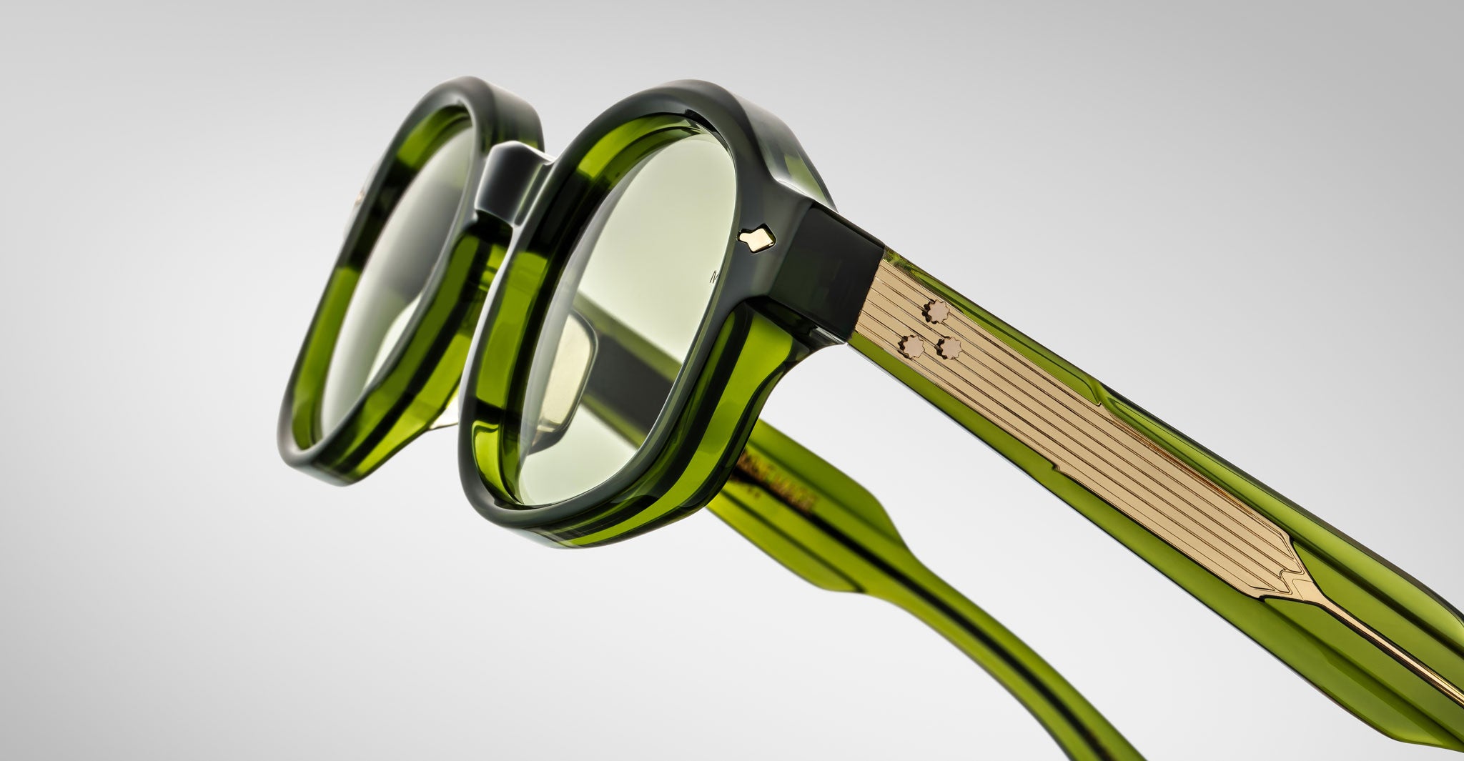 A close-up of Jacques Marie Mage Petrus green translucent round eyeglasses with gold metallic arm detailing, set against a neutral gradient background.