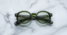 A pair of Jacques Marie Mage Petrus round green eyeglasses with thick frames and slightly green-tinted lenses rests on a white marble surface, their arms neatly folded.