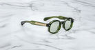 A pair of Jacques Marie Mage Petrus round green sunglasses with gold temple detailing rests on a white marble surface.