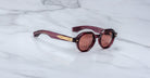 The Jacques Marie Mage Pontian, round dark red sunglasses with gold temple accents, is displayed on a white marble surface.