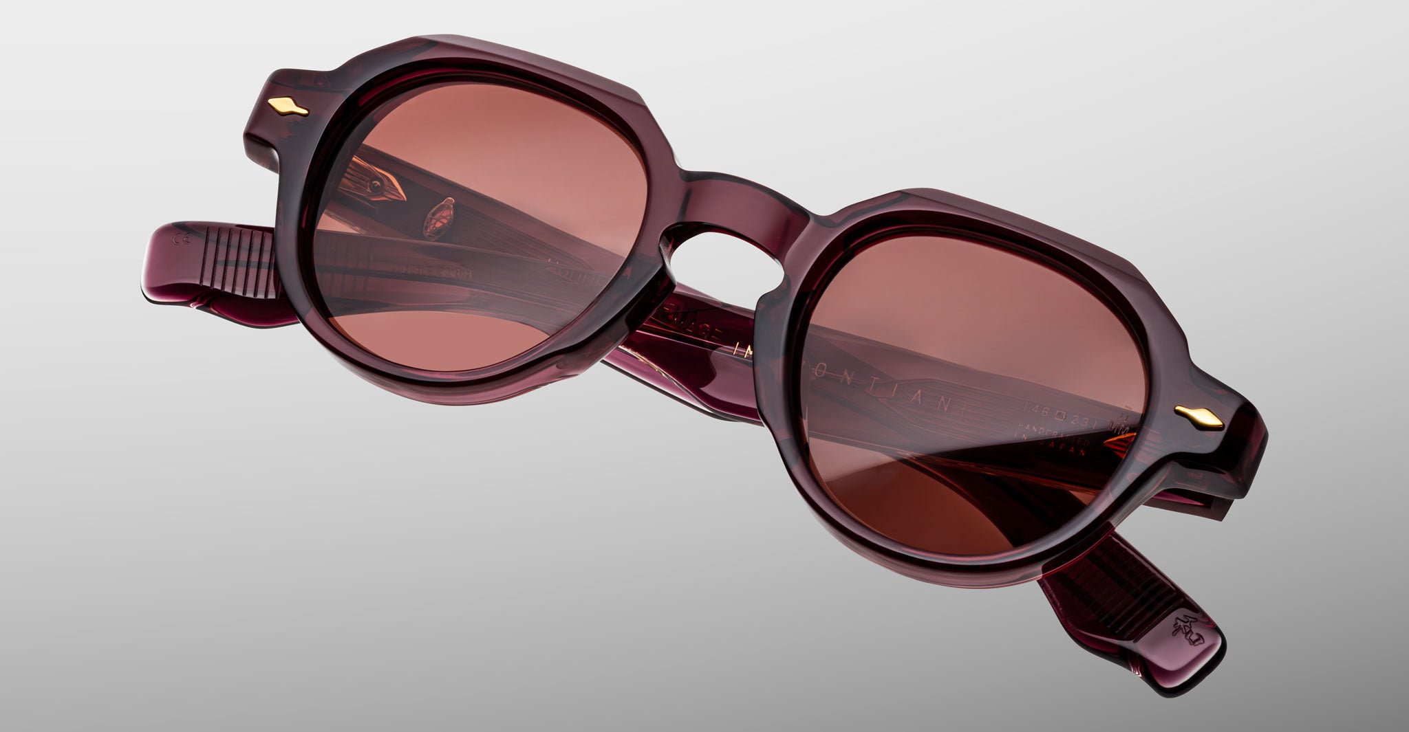 The Jacques Marie Mage Pontian sunglasses feature round, thick, glossy burgundy frames and reddish-brown tinted lenses, showcased against a light gray background.