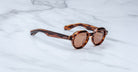 A pair of Jacques Marie Mage Pontian round, brown tortoiseshell sunglasses with dark lenses rests on a white marble surface.