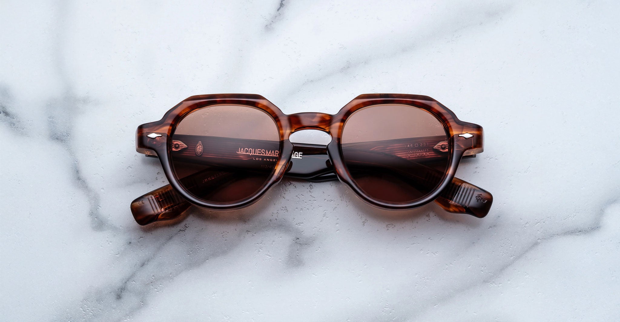 The Jacques Marie Mage Pontian sunglasses, featuring round, slightly geometric brown-tinted lenses and thick arms in a brown tortoiseshell design, are displayed on a white marble surface.