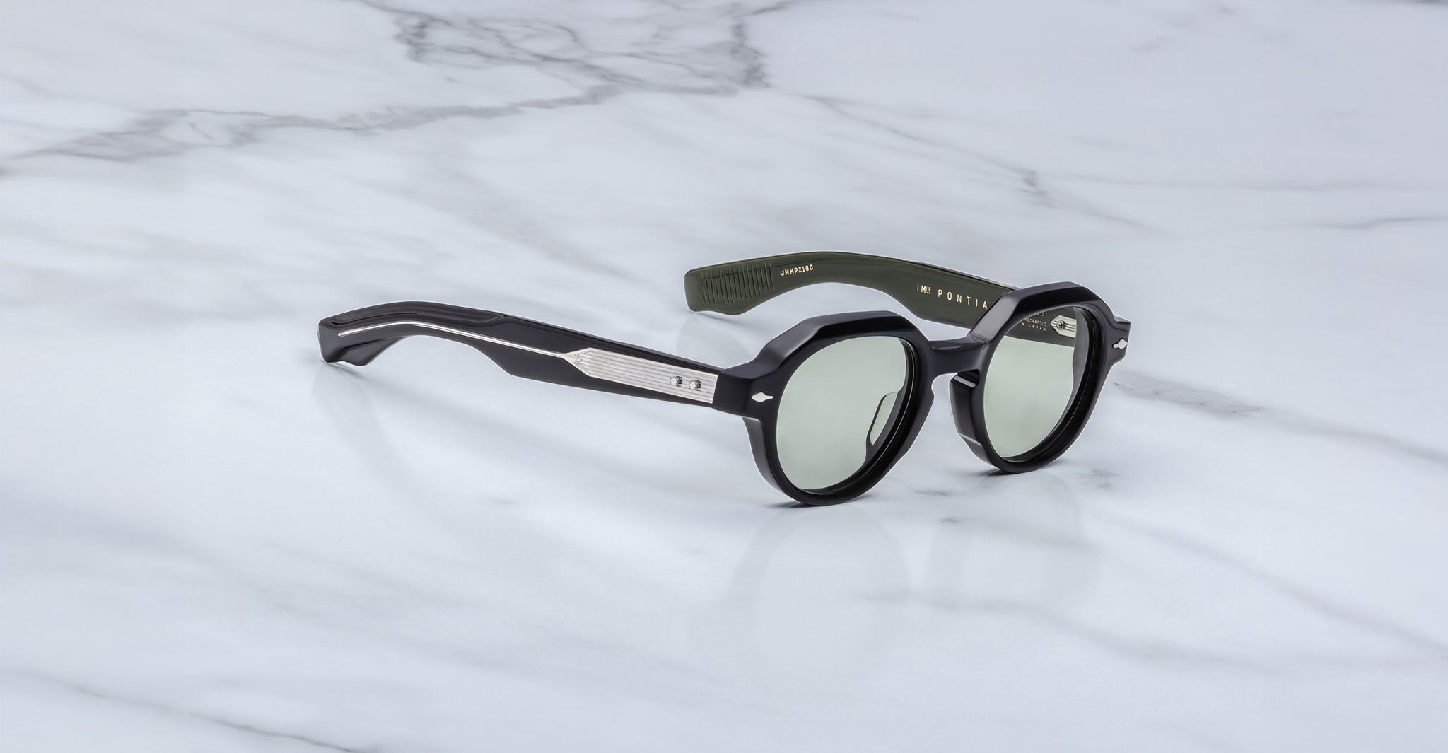 A pair of Jacques Marie Mage Pontian round, black sunglasses with silver detailing on the arms rests on a white marble surface. The tinted lenses and branded text inside the arms add a touch of sophistication.