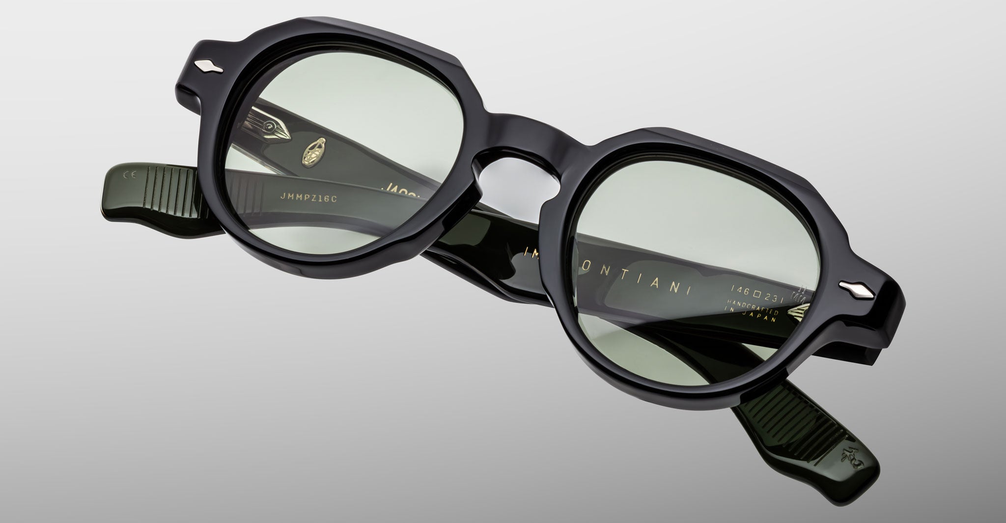 The Jacques Marie Mage Pontian sunglasses in black, featuring geometric frames and green-tinted lenses, are displayed on a plain light gray background. The brand name and details appear on the inside of the arms.