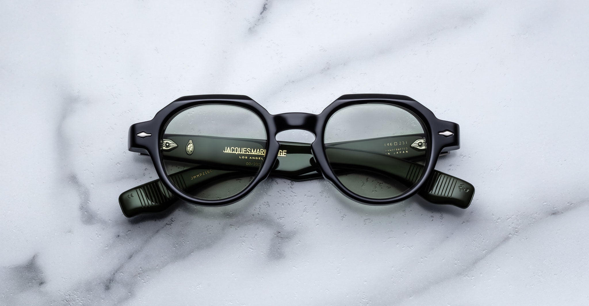 The Jacques Marie Mage Pontian is a pair of black, round eyeglasses with thick frames, shown lens-down on a white marble surface; their folded arms are visible through the lenses.