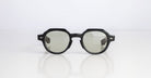 The Jacques Marie Mage Pontian sunglasses, featuring thick geometric black frames and gray tinted lenses, are centered on a reflective white surface against a plain light-colored background.